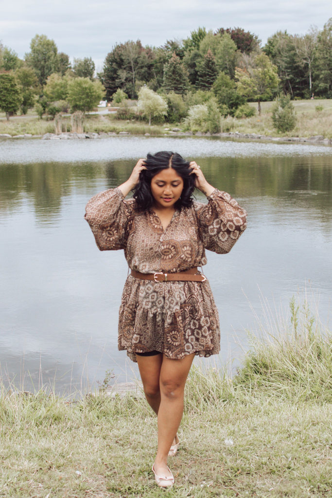 Canadian lifestyle blogger, Lisa Favre, wearing a light, airy brown dress, with curly short hair. Posing in a park in front of a pond.