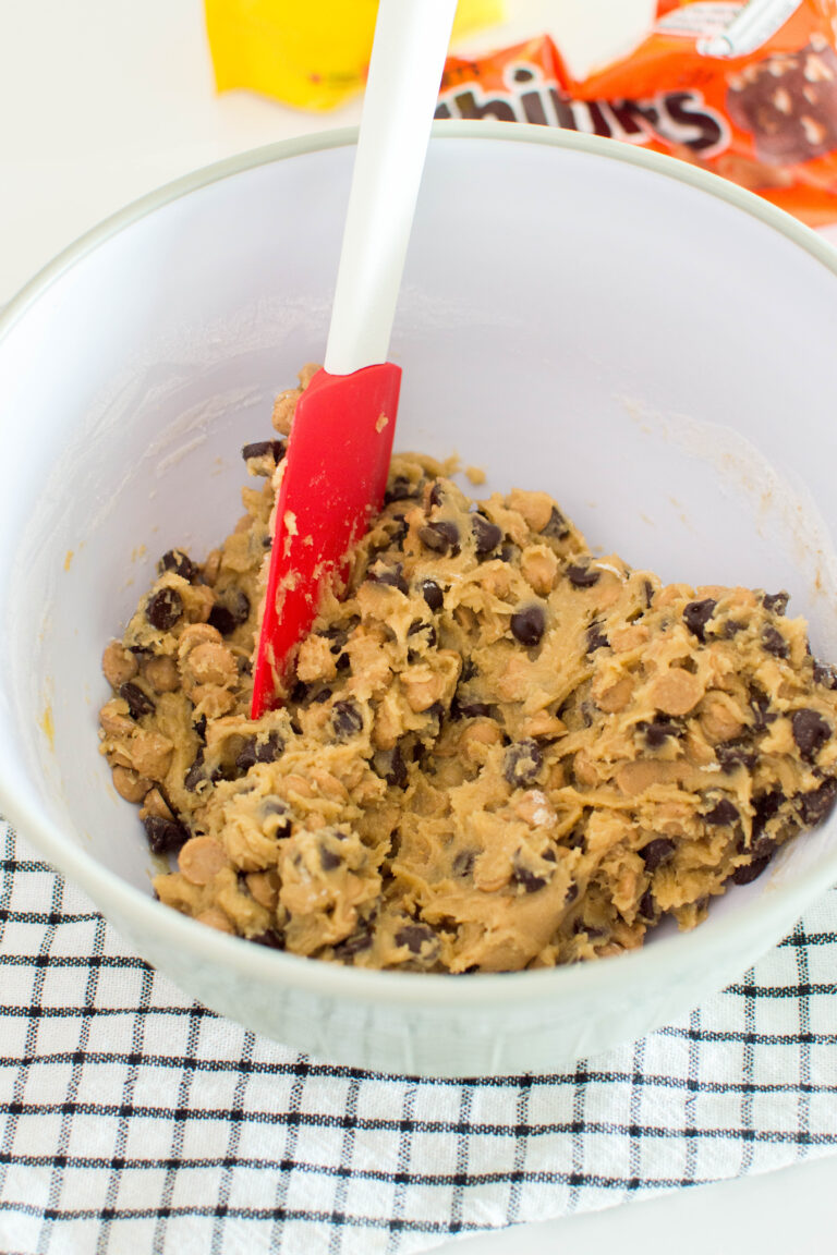 Adding chocolate chips to a bowl of cookie dough, being mixed with a red spatula