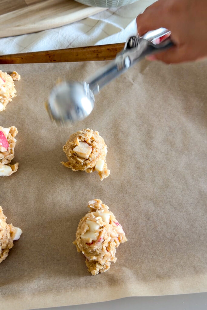 Scooping apple crumble cookie dough onto a parchment-lined baking sheet.