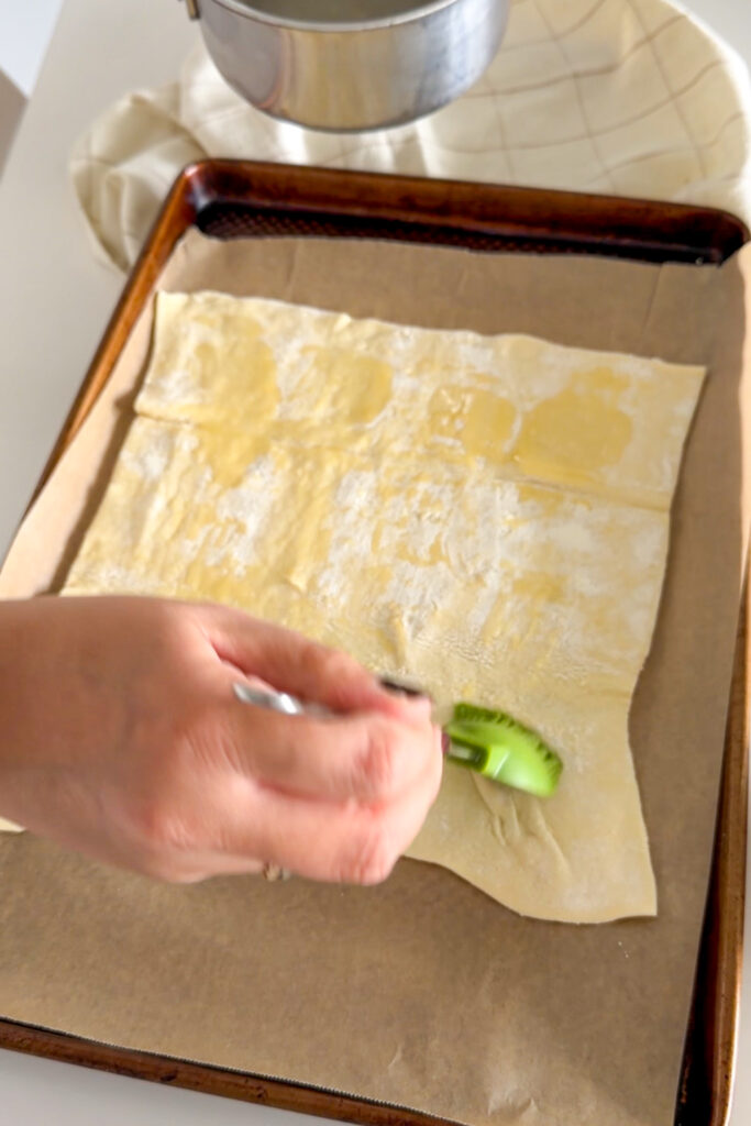 Brushing a sheet of puff pastry with melted butter on a parchment-lined baking sheet.