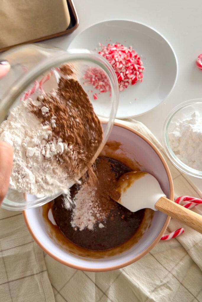Adding dry ingredients to wet ingredients to make Peppermint Chocolate Crinkle Cookies with a plate of crushed candy canes on the side